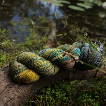 A Worsted yarn skein colored with green, yellow and grey colors, with blue speckles, on top of a tree limb that is hovering over a pond, with different mossy plants around the pond, and around the yarn.