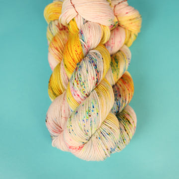 A light cream colored Worsted yarn with different colored speckles all over and banana colored yellow at the top of the skein on top of a light blue background.