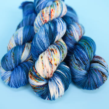 Worsted yarn colored with different shades of blue and white with multicolored speckles on the portion of the yarn that Is white. all on top of a light blue background.