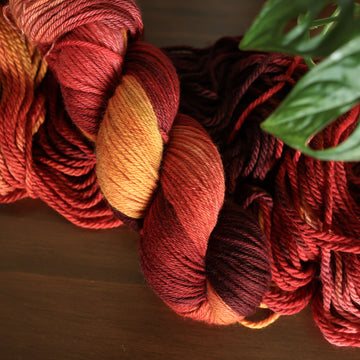 Worsted yarn with red orange and yellow colors, on top of an unwound version of that same yarn. all on top of a dark wooden table with a couple of leaves of a plant overhead.