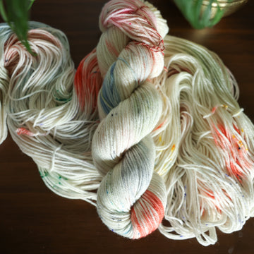 worsted white yarn with mixed colors of light pink purple and red/orange and brown with speckles, on top of an unwound version of that same yarn. all on top of a dark wooden table with a couple of leaves of a plant overhead in a gold pot.