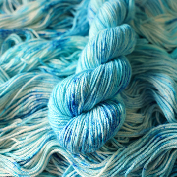 A DK skein of yarn colored bright and frosty blue with cobalt speckles. The skein is sitting on the unwrapped yarn.