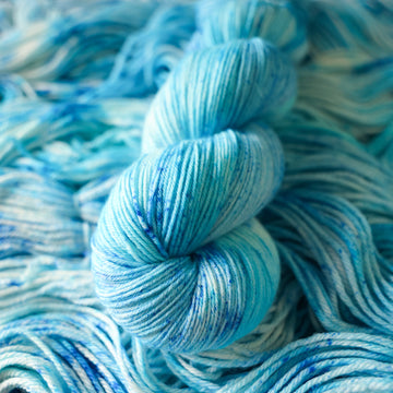 A fingering skein of yarn colored bright and frosty blue with cobalt speckles. The skein is sitting on the unwrapped yarn.