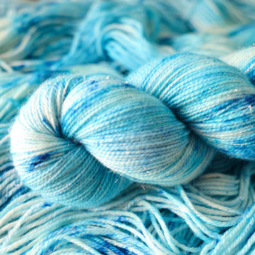 A sparkle skein of yarn colored bright and frosty blue with cobalt speckles. The skein is sitting on the unwrapped yarn.