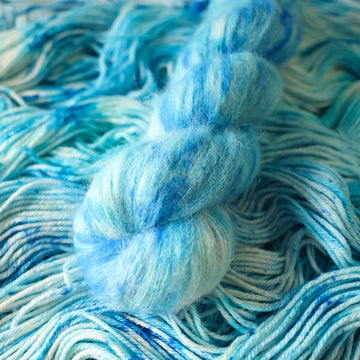 A suri skein of yarn colored bright and frosty blue with cobalt speckles. The skein is sitting on the unwrapped yarn.