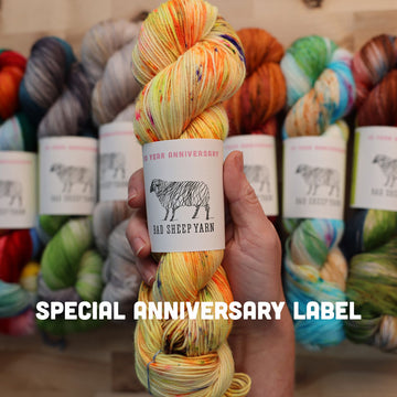 Different color ways of wrapped yarn lined up next to each other on a wooden table, with someone holding a yellow/orange yarn that is wrapped up, with the words special anniversary label at the bottom of the picture.