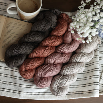 Four DK skeins of yarn sitting side by side each other. From left to right, one skein is colored brown tonal solid with green undertones,  beside it is  a colored moody dark salmon pink tonal solid, next is a colored a moody dark salmon pink tonal solid, last is a a misty morning grey tonal solid. The first skein his sitting on top of a book and next to it is a mug of coffee. In the corner there is a blue and white vase with white flowers. All sitting on top of a black and white striped table cloth.