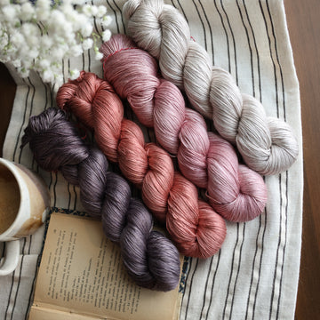 Four silk skeins of yarn sitting side by side each other. From left to right, one skein is colored brown tonal solid with green undertones,  beside it is  a colored moody dark salmon pink tonal solid, next is a colored a moody dark salmon pink tonal solid, last is a a misty morning grey tonal solid. The first skein his sitting on top of a book and next to it is a mug of coffee. In the corner there is white flowers peeking out. All sitting on top of a black and white striped table cloth.