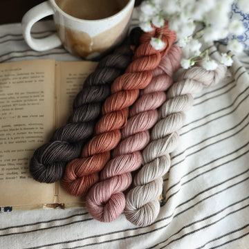 Four sock mini skeins of yarn sitting side by side each other. From left to right, one skein is colored brown tonal solid with green undertones,  beside it is  a colored moody dark salmon pink tonal solid, next is a colored a moody dark salmon pink tonal solid, last is a a misty morning grey tonal solid. The first skein his sitting on top of a book and next to it is a mug of coffee. In the corner there is a blue and white vase with white flowers. All sitting on top of a black and white striped table cloth.
