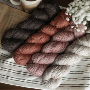 Four Sparkle sock skeins of yarn sitting side by side each other. From left to right, one skein is colored brown tonal solid with green undertones,  beside it is  a colored moody dark salmon pink tonal solid, next is a colored a moody dark salmon pink tonal solid, last is a a misty morning grey tonal solid. The first skein his sitting on top of a book and next to it is a mug of coffee. In the corner there is white flowers peeking out. All sitting on top of a black and white striped table cloth.