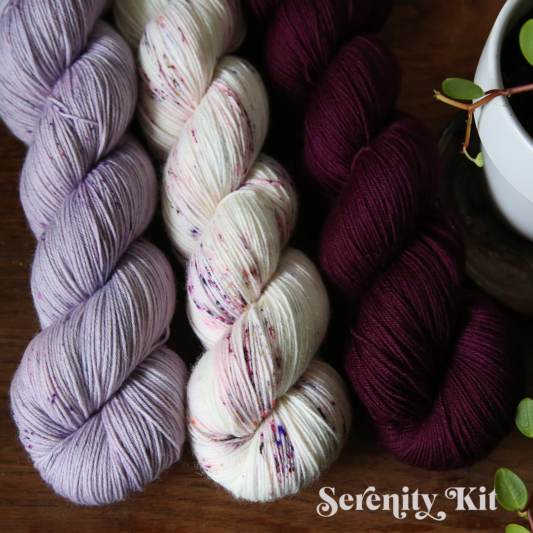 three skeins of yarn are arranged on a wooden surface in lavender, speckled white, and deep burgundy colors