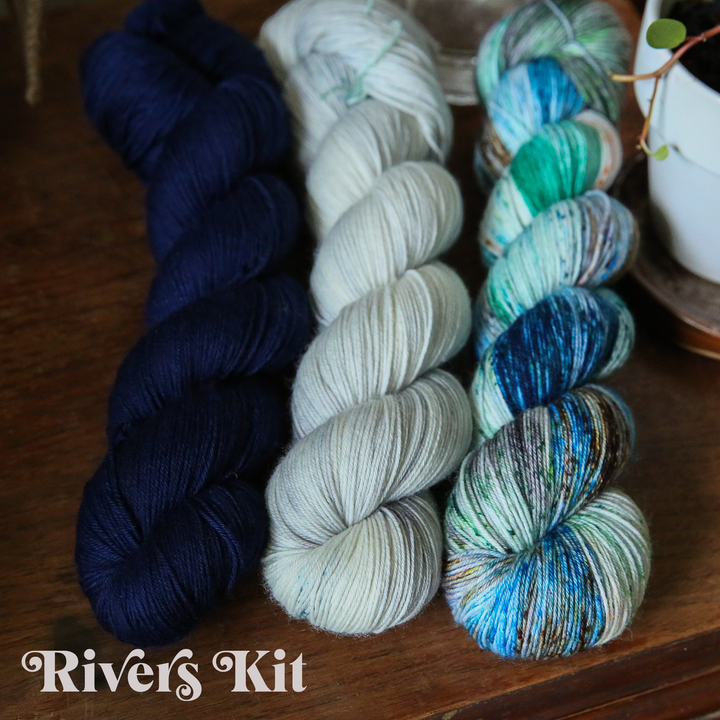 yarn skeins in solid navy blue, solid off-white, and a variegated blue, green, and teal on a wooden surface